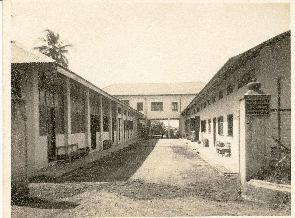 Old black and white photo of a dirt courtyard flanked by long low buildings with a two-story structure at the far end.